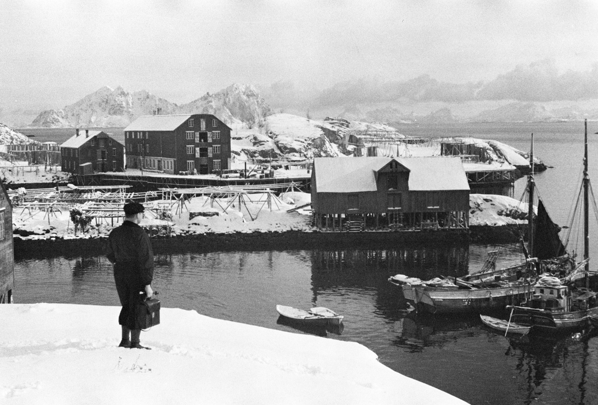 Anders Beer Wilse, Mann med dokumentmappe står avventende i sjøkanten, fiskebåter og naust langs bryggene.