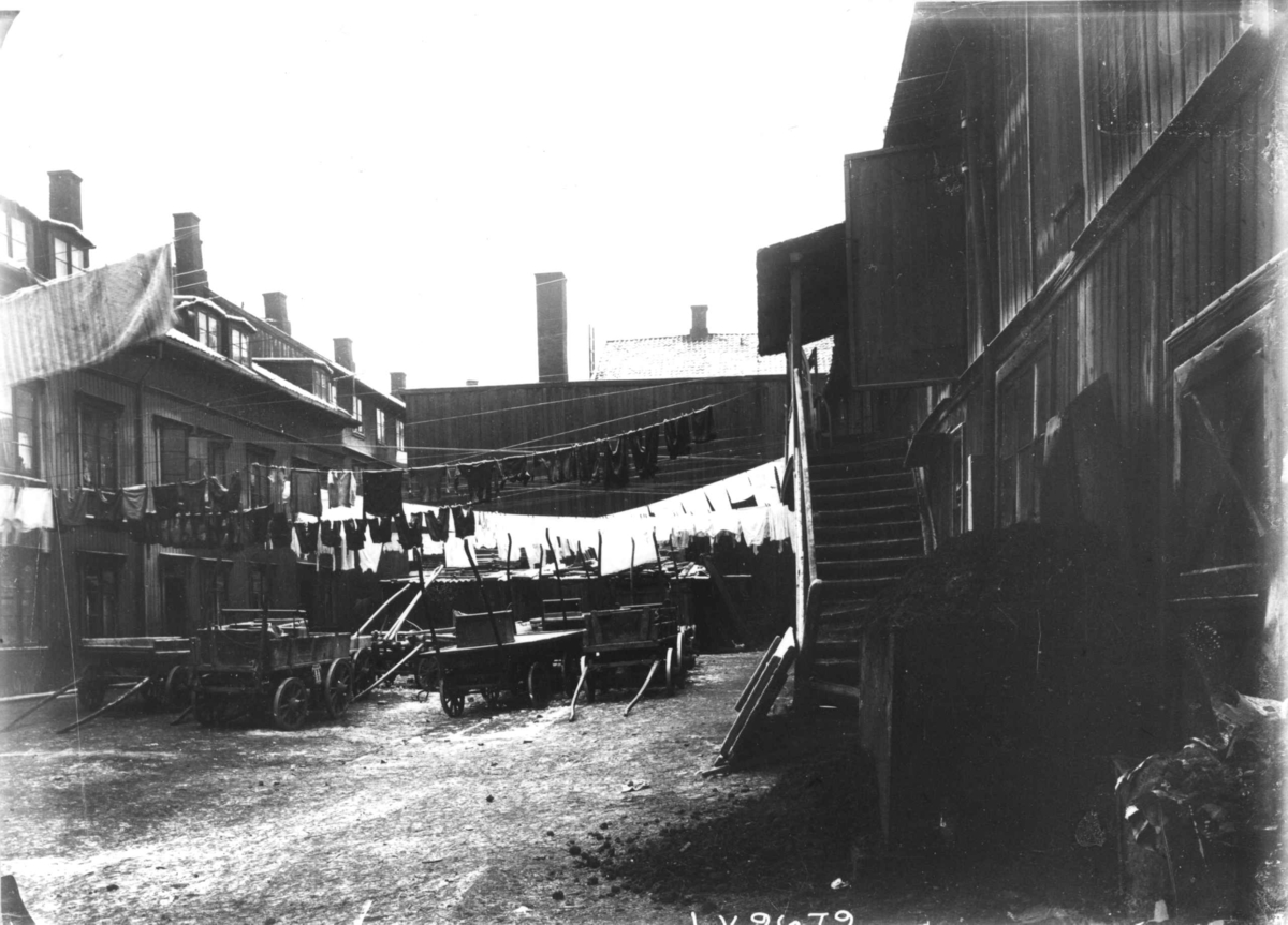 Nanna Broch, Gårdsplass med boliger, muligens på Rodeløkka, Oslo. Hestekjøretøyer plassert på plassen med klesvask over. Fra boliginspektør Nanna Brochs boligundersøkelser i Oslo 1920-årene.