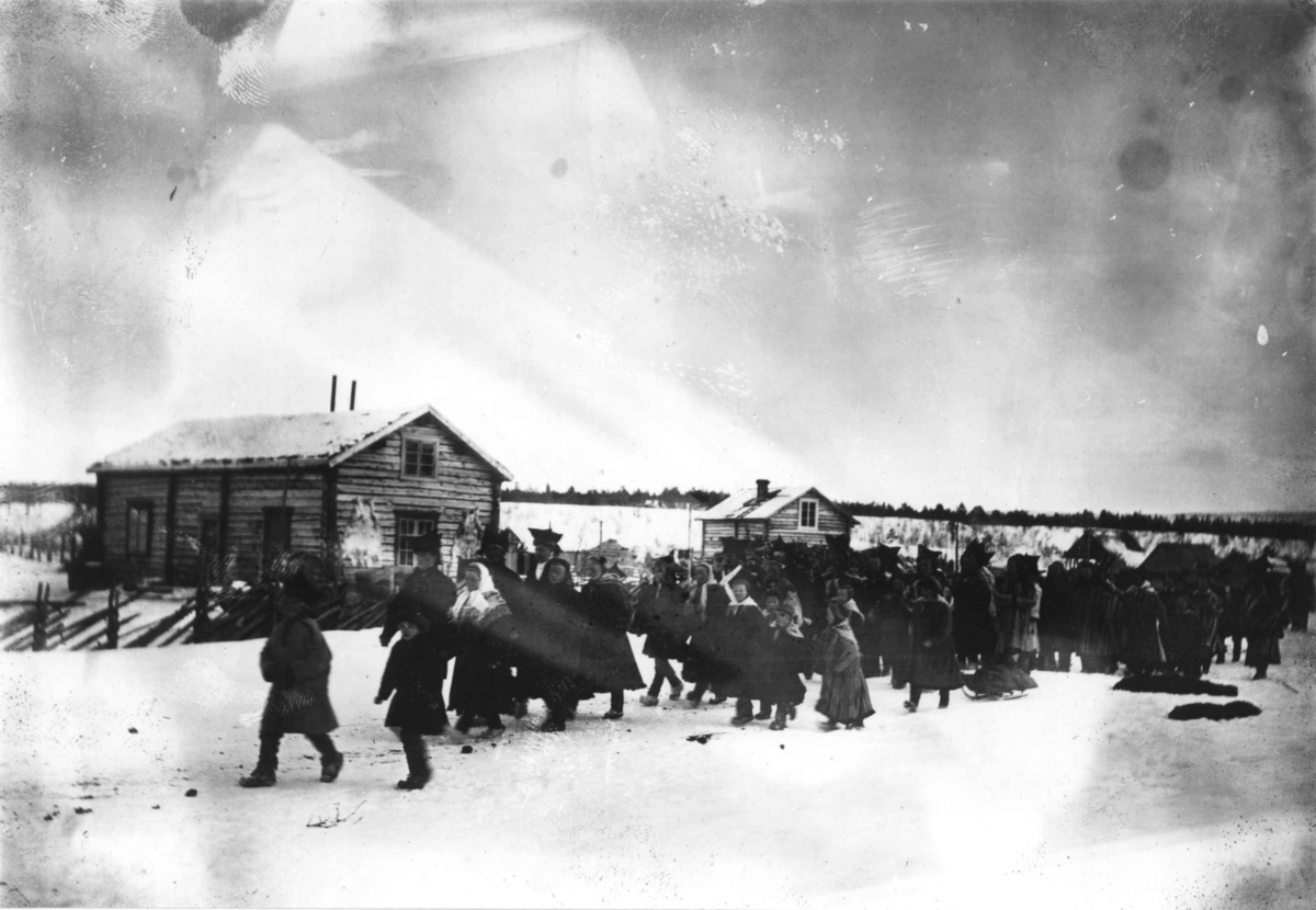Konrad Nielsen, Brudefølge i Karasjok, brudgommen med hvitt bånd og bruden hvitt sjal. To hus i bakgrunnen.