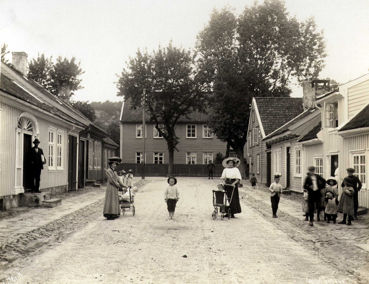 Anders Beer Wilse, Bygate med beboere, Kristiansand, Vest-Agder. Barn og voksne i trehusbebyggelse. Kvinner med barnevogner.
