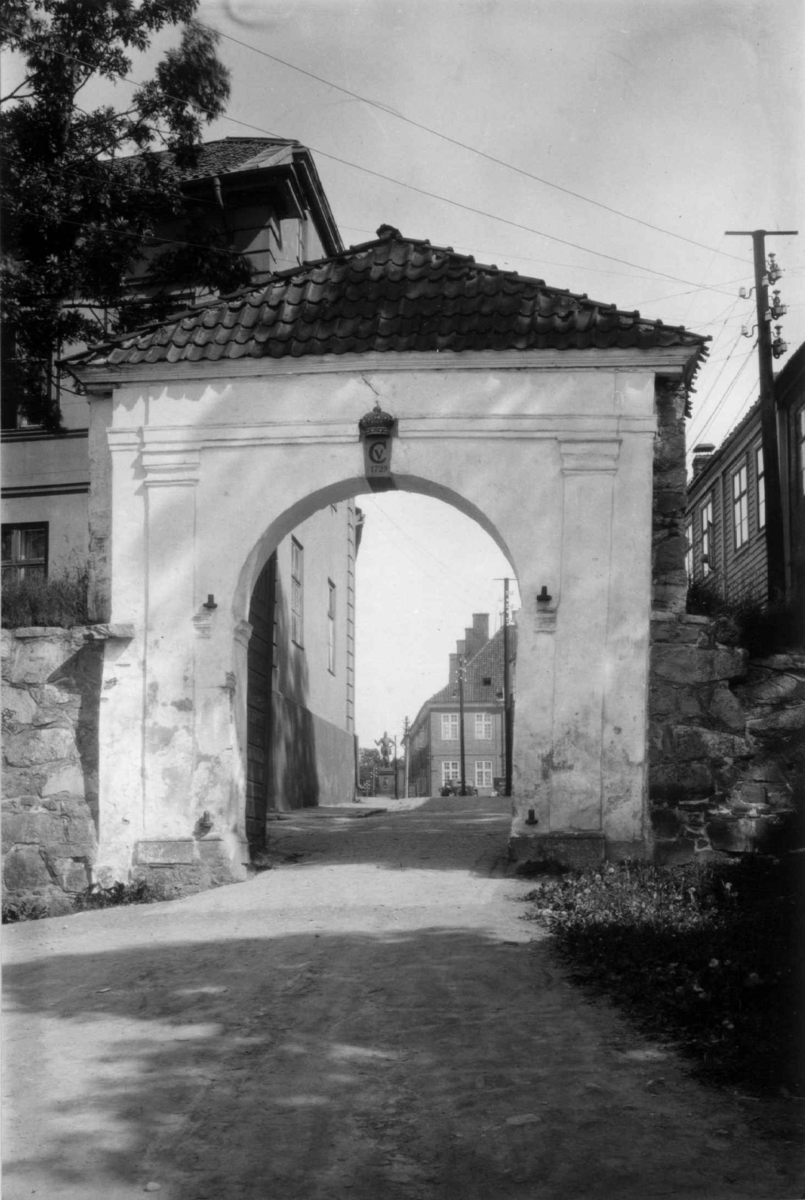 Hermann Christian Neupert, Fredrikstad. 1930. Byport med initialene til Christian V og årstallet 1729. Gate og bebyggelse i bakgrunnen.