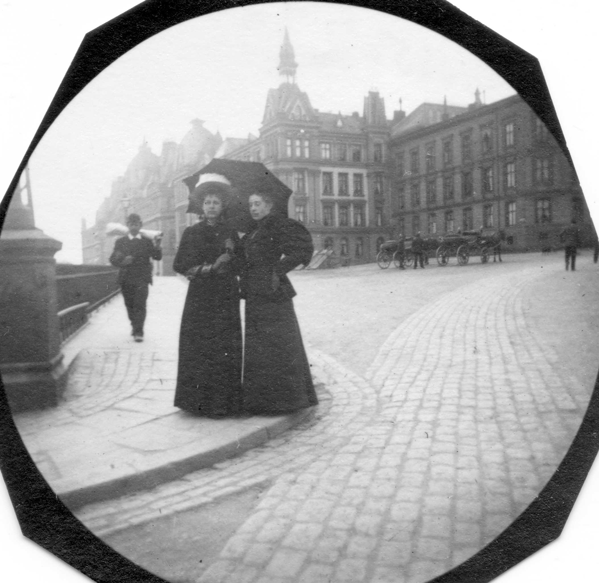 Fredrik Carl Mülertz Størmer, Thora Christoffersen og Alette Thoulow, senere fru Fougner, står på fortau ved Victoria terrasse i Oslo. Plass med bygårder og hestedrosjer.