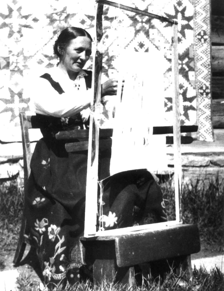 Anna Grostøl, Antagelig Lesja, Oppland 1934. Kvinne i arbeid ved sprangramme. et teppe henger på husveggen i bakgrunnen.