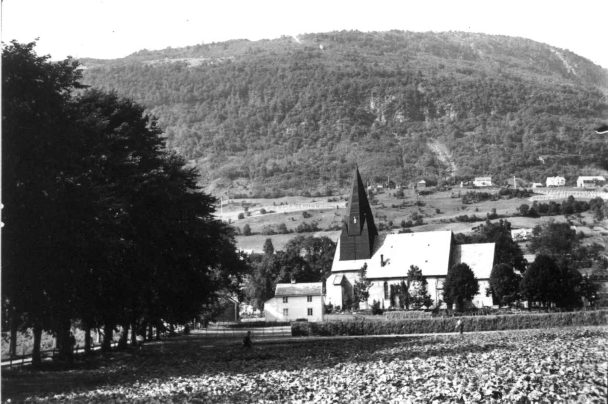 Hermann Christian Neupert, Voss kirke 1939. Landskap med gårder, skog og fjell i bakgrunnen. I forgrunnen en allé og et jorde med hesje.