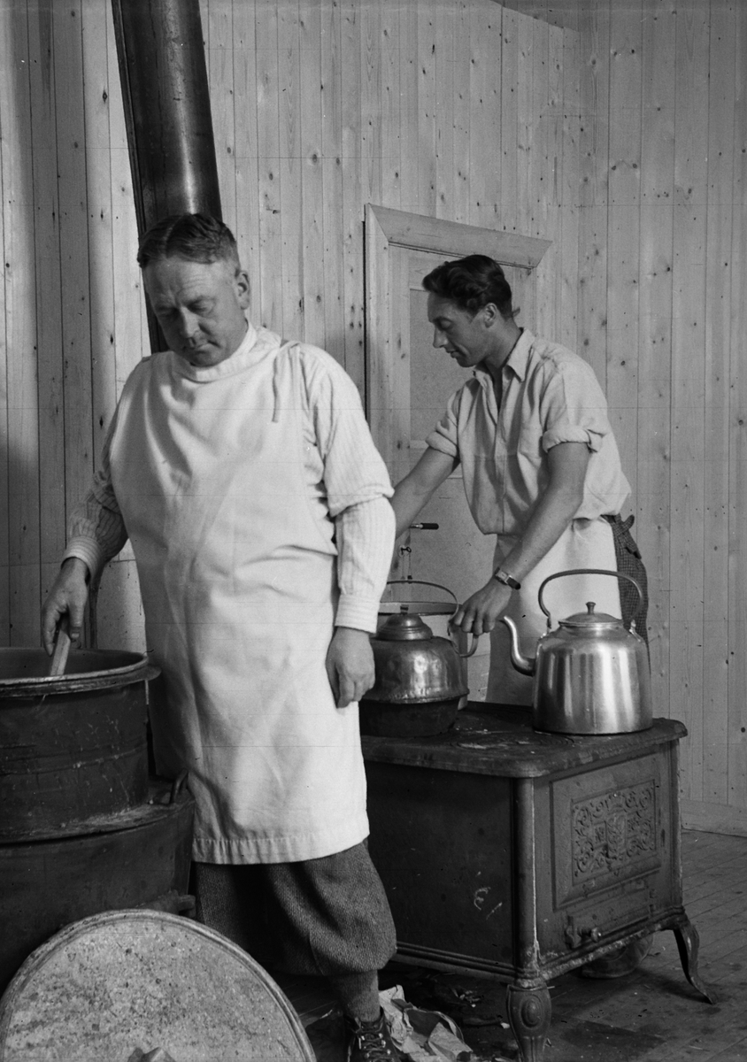 Anno Domkirkeodden, INTR: KJØKKEN, VEDKOMFYR, BRYGGERPANNE, 2 MENN M/FORKLE LAGER MAT LØTEN. MATLAGING.