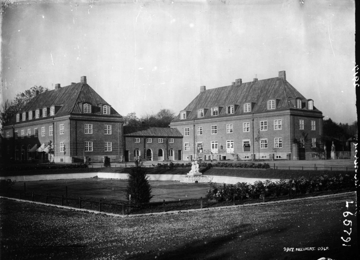 Hermann Christian Neupert, Damplassen, Ullevål Hageby, Oslo. 1925. Bebyggelse med forretninger i bakgrunnen. Beplantning i forgrunnen.