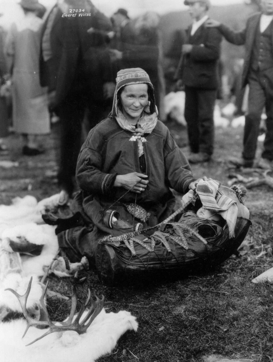 Anders Beer Wilse, En kvinne sitter ved et barn i komse, mennesker i bakgrunnen. Antagelig Lyngen 1925.