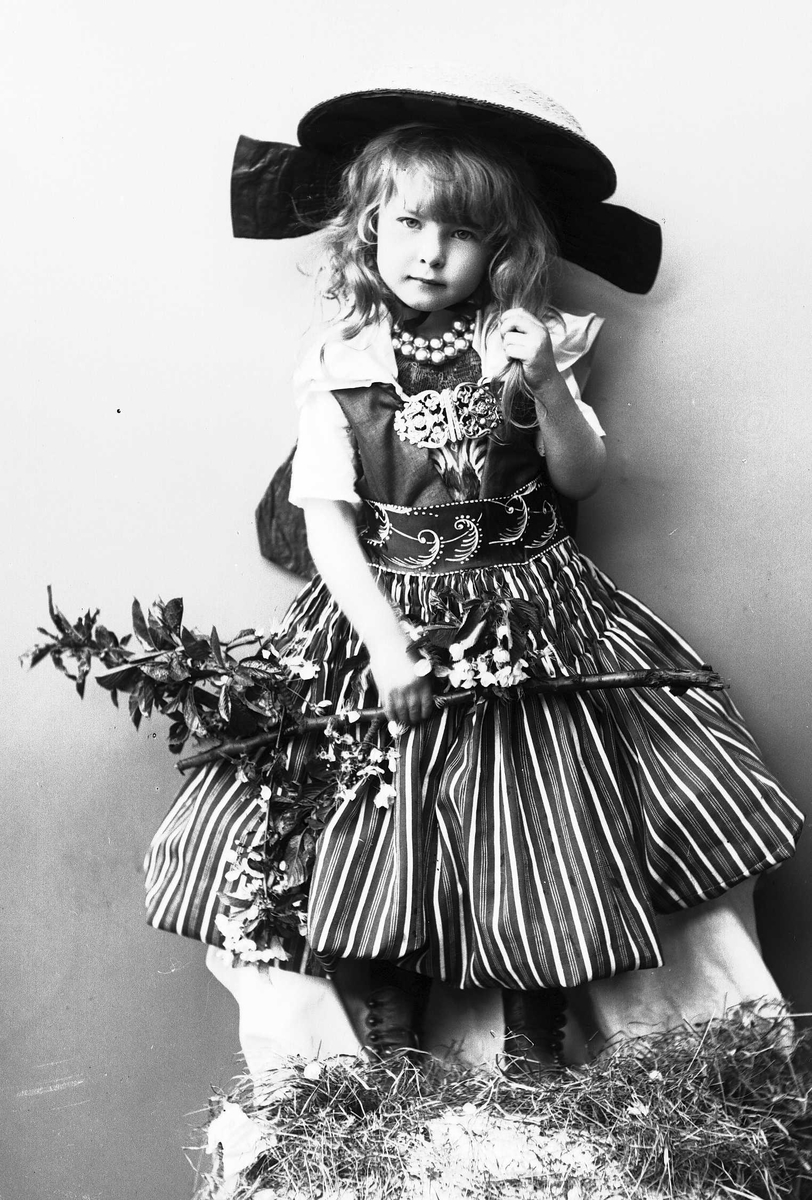 Solveig Lund, Pike i drakt og hatt, med perlekjede i halsen. Holder gren med blader og blomster.