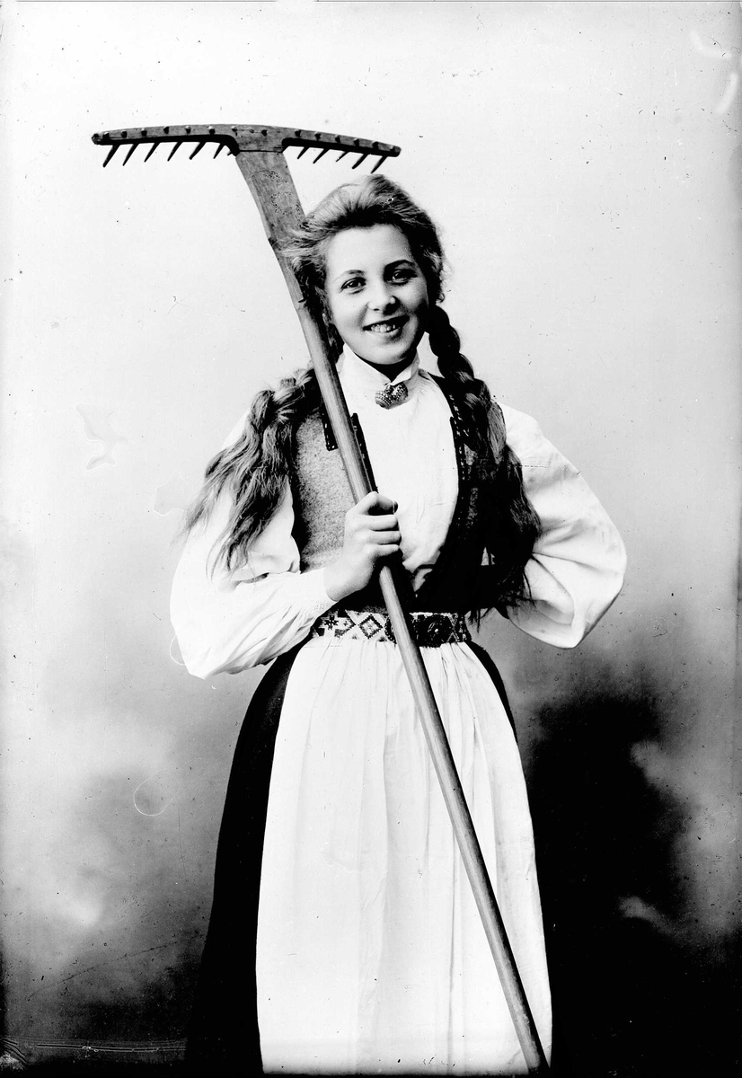 Solveig Lund, 3/4 dels studioportrett av kvinne i drakt, med flettet hår. Hun holder en rive i høyre arm, mens den venstre hviler mot hoften. Bakgrunnen er nøytral.