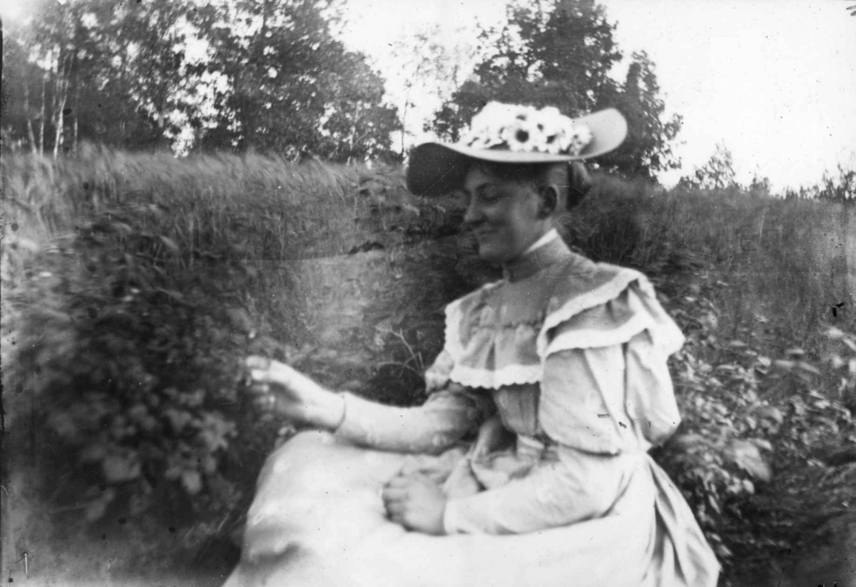Hermann Christian Neupert, Drakt 1908-10. Kvinne med kjole og hatt sittende i en hage(?).