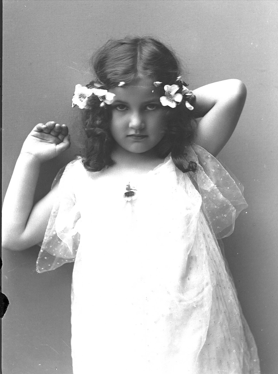 Solveig Lund, 3/4 portrett av jente med kjole og blomster i håret. Løfter hendene bak hodet og ser på betrakteren.