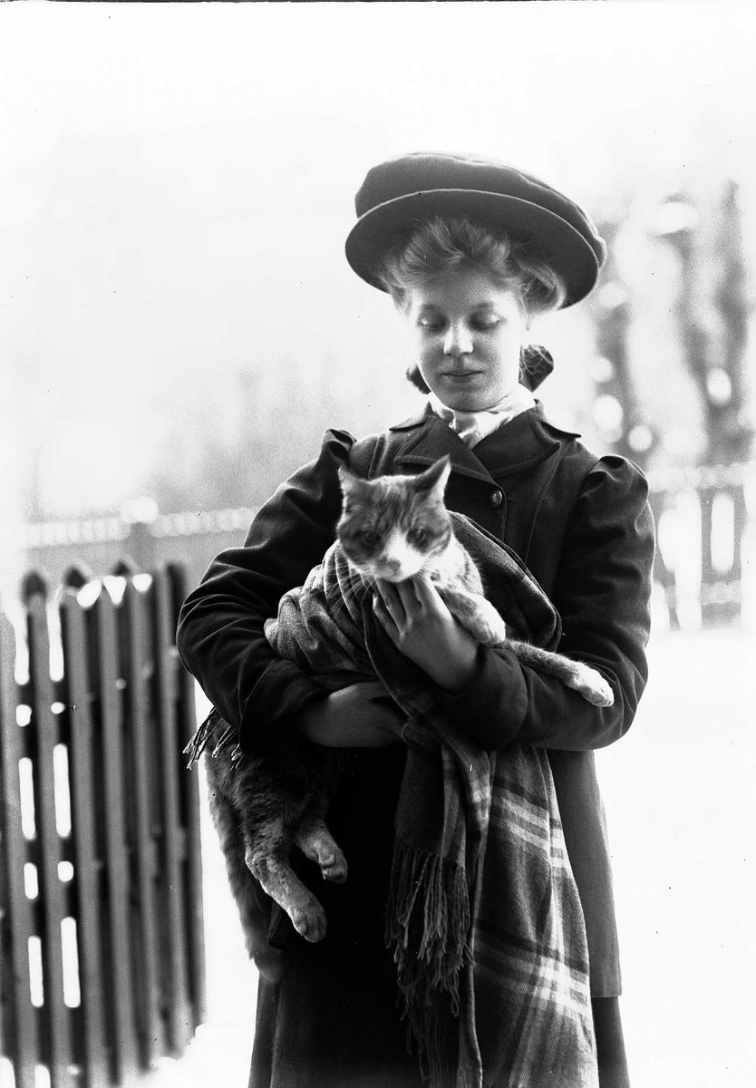 Solveig Lund, Utendørsfotografi av kvinne i kåpe og hatt, som holder katt under ullpledd.