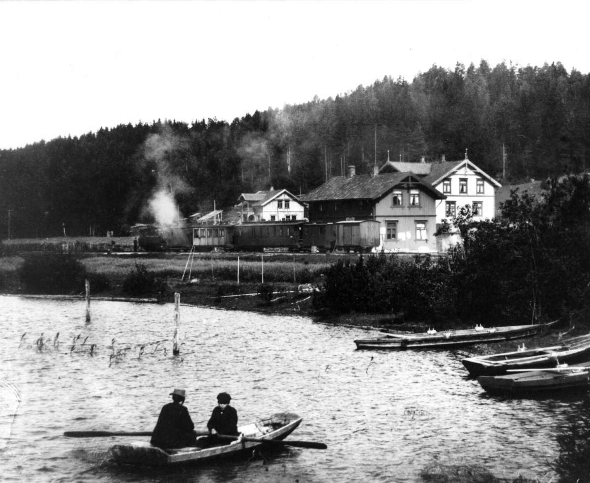Anders Beer Wilse, Skullerud, Aurskog-Høland, Akershus 1904. To mennesker i robåt på vann foran jernbanestasjon. Urskog-Hølandsbanen.