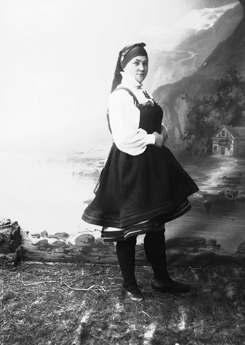 Solveig Lund, Studioportrett av kvinne i nasjonaldrakt avbildet fra siden. Hun står vendt mot høyre og ser ut mot betrakteren. Står på gress/jordbunn. Malt bakgrunn med fjord og fjell og et lite hus.