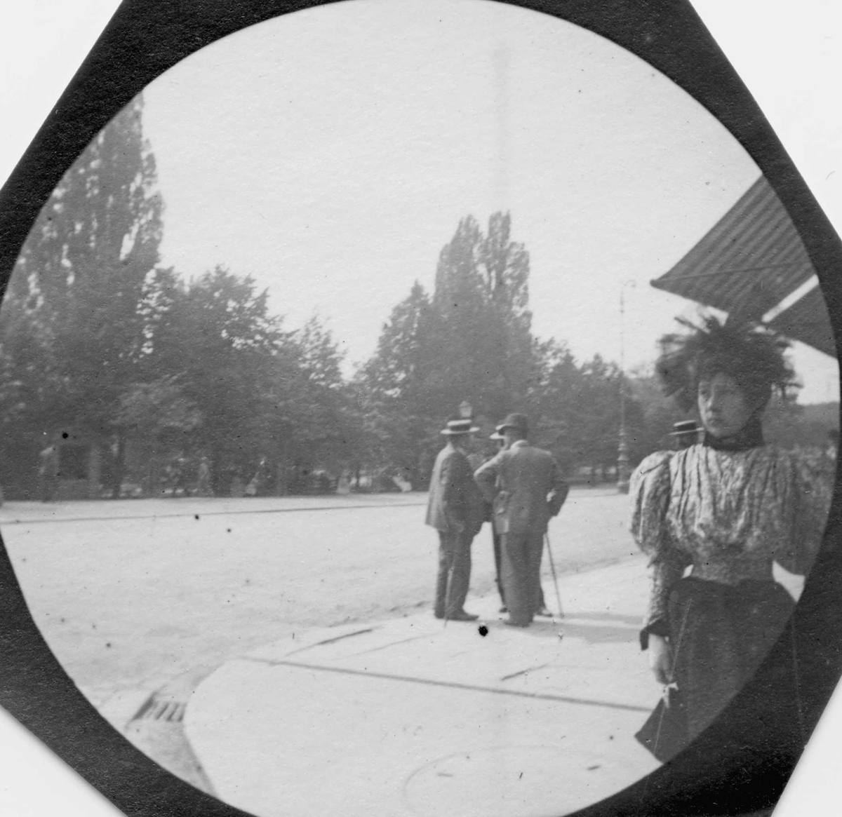 Fredrik Carl Mülertz Størmer, Kvinne spaserer langs bygate, ant. Karl Johans gate, Oslo.. Tre menn står og prater. Park på den andre siden.