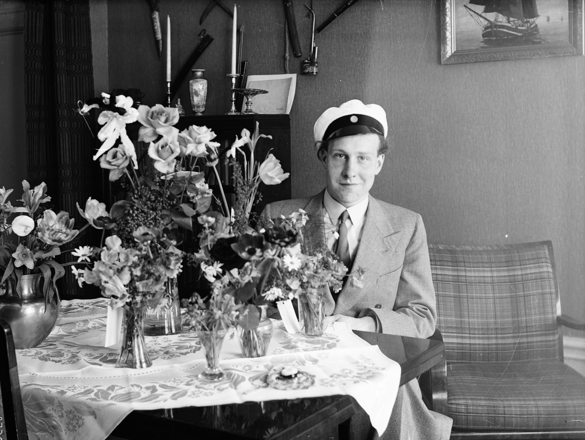 Okänd,, Studentfirande, ung man sitter vid bord med blommor, sannolikt hemma i Sverige