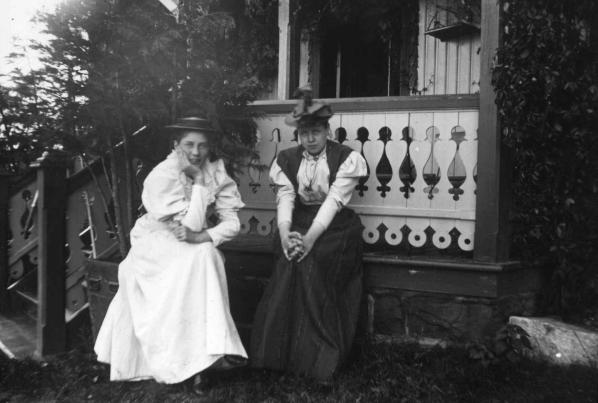 Hermann Christian Neupert, Div. drakt, Norge. 1908-1910.To kvinner sittende i en hage. En kledd i lang kjole og en i langt skjørt og bluse. Hatter på hodet. Veranda i bakgrunnen.