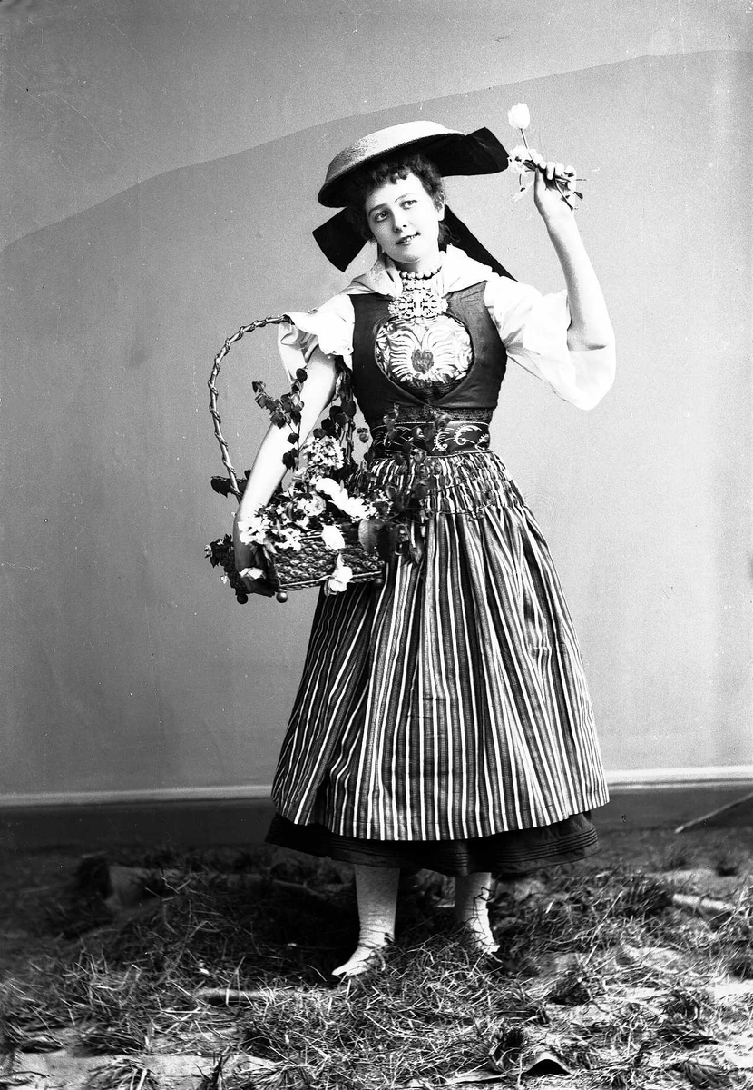 Solveig Lund, Kvinne i drakt og hatt, som holder kurv med blader og blomster. Står på høy.