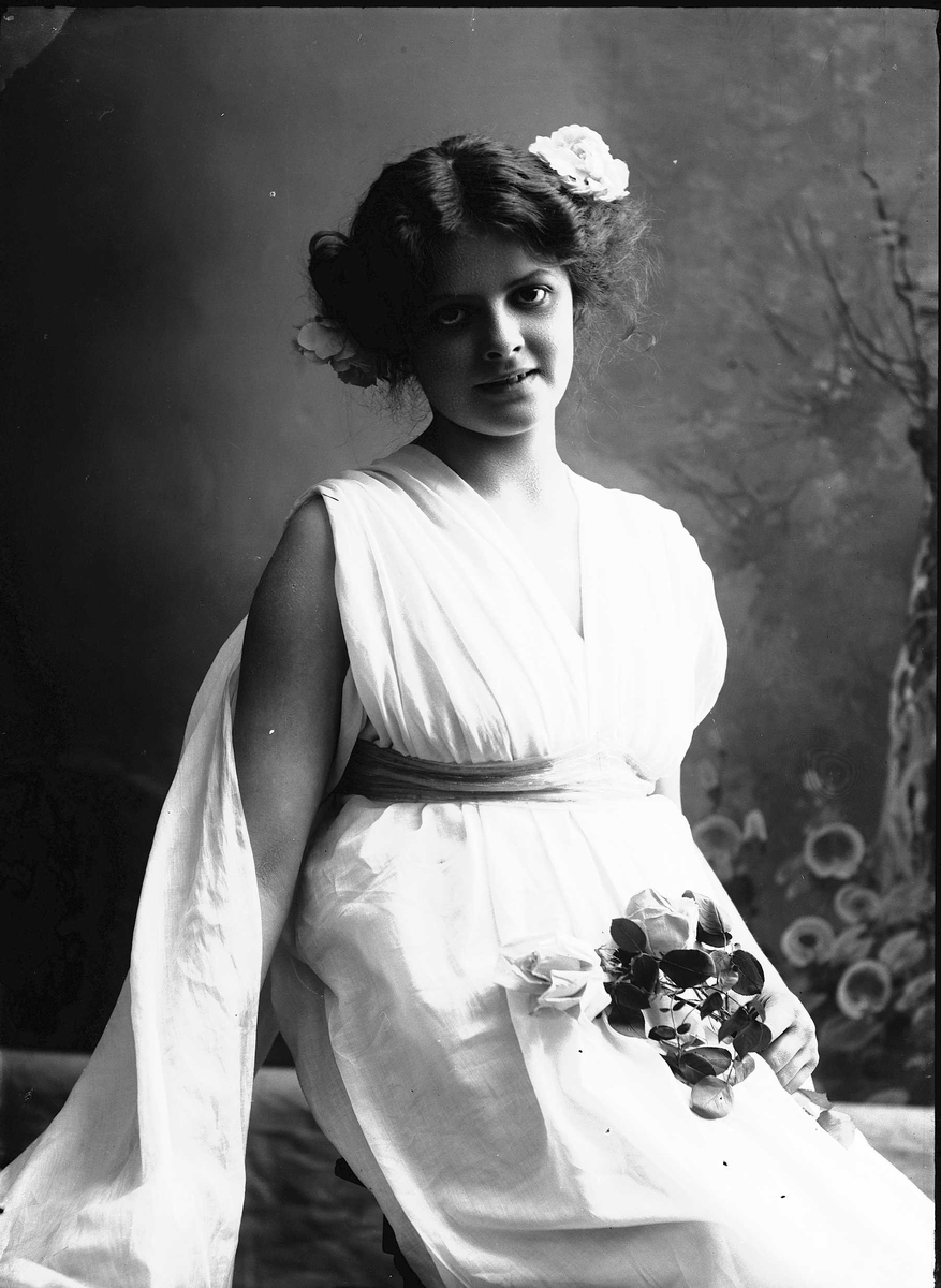Solveig Lund, Studioportrett av kvinne i kostyme med hvit kjortel og blomster i håret. I den ene hånden holder hun en blomsterbukett og bakgrunnen er malt med trær og blomster.