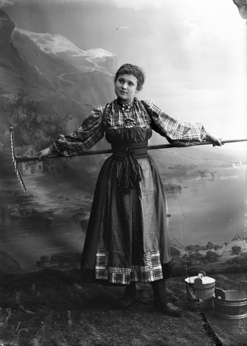 Solveig Lund, Studioportrett av kvinne i nasjonaldrakt som holder rive bak ryggen. To tiner står på bakken til venstre for henne. Bakgrunnen er malt med fjell, fjord og hus. På bakken er det gress.
