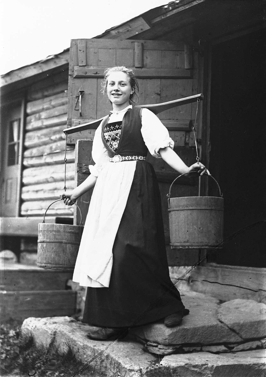 Solveig Lund, Pike i Hardangerdrakt står foran dør til seter. Bærer på to vannbøtter.