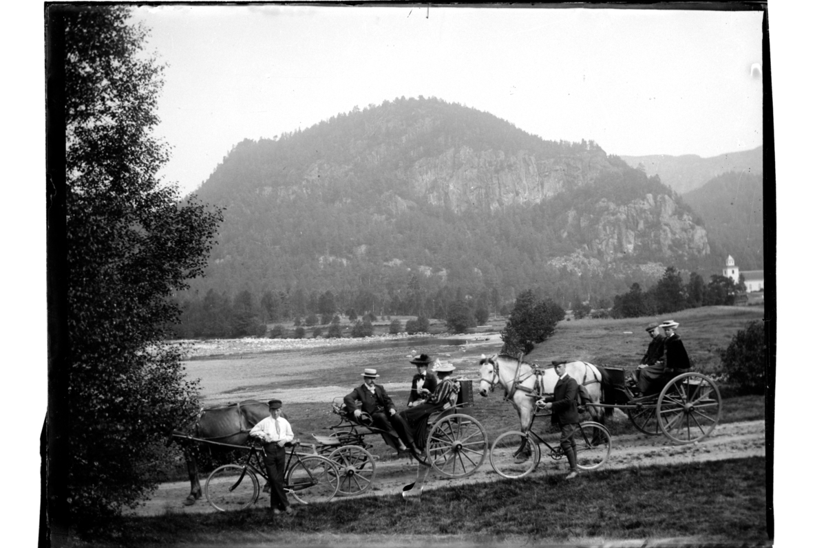 Rolf Sr. Sundt, To hestekjøretøy med passasjerer og to syklister i ukjent landskap med kirke. Fotografert rundt 1902.
