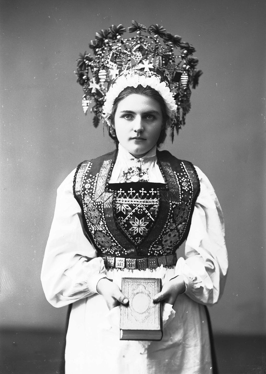 Solveig Lund, Studiofotografi av kvinne med brudedrakt og brudekrone fra Voss i Hordaland. Holder på salmebok/bibel. Stående. Hofteutsnitt. Frontalt vendt. Ser ut av bildet.