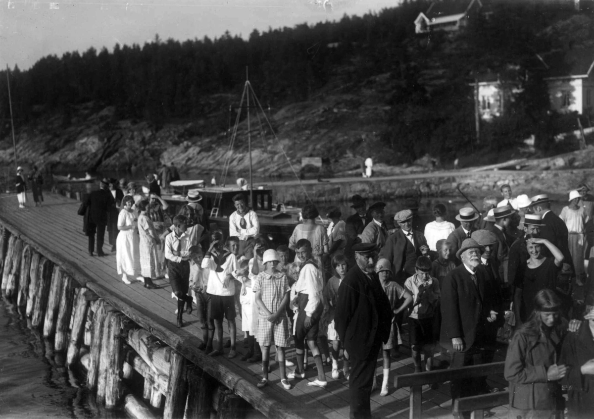 Hermann Christian Neupert, Hvitsten, Vestby, Akershus 1924. Oversiktsbilde. Festkledde mennesker samlet på brygge. Hus i bakgrunnen.
