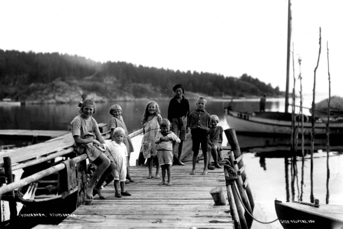 Hermann Christian Neupert, Barneklær. Frogn, Drøbak, Akershus. 1924. Barn i sommerklær på en brygge. Jentene i kjoler. Guttene i shorts. En liten gutt i matrosdress. Fjorden i bakgrunnen. Båter.