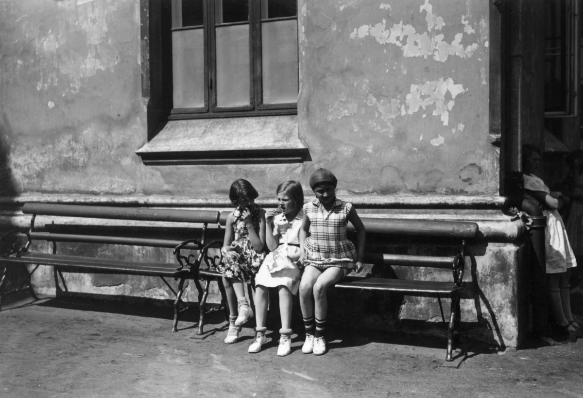 Hermann Christian Neupert, Oslo. Småjenter kledd i kjoler på en benk ved Ruseløkka skole. Mai 1932.