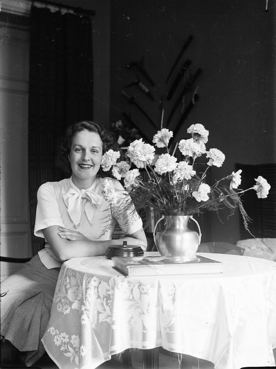 Okänd,, Ung kvinna sitter vid bord med blommor 1930-40-tal, sannolikt i hemmet , Sverige