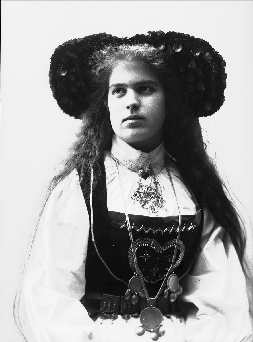 Solveig Lund, Studiofotografi av kvinne med brudedrakt og brudehatt på hodet. Sittende. Hofteutsnitt. Vendt mot høyre. Ser ut mot venstre.