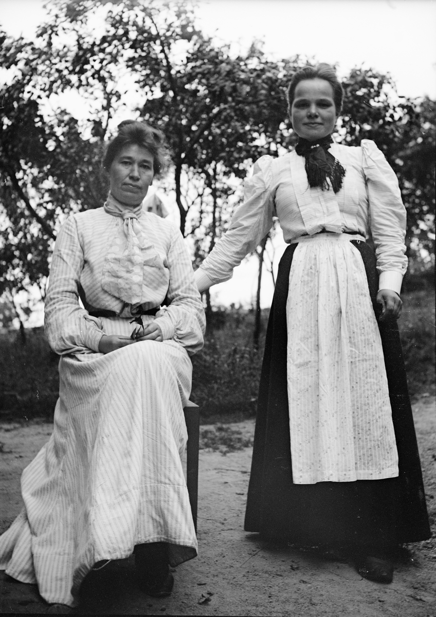 Okänd,, Två kvinnor, en sitter och den andra står, ute i trädgård, sannolikt i Sverige