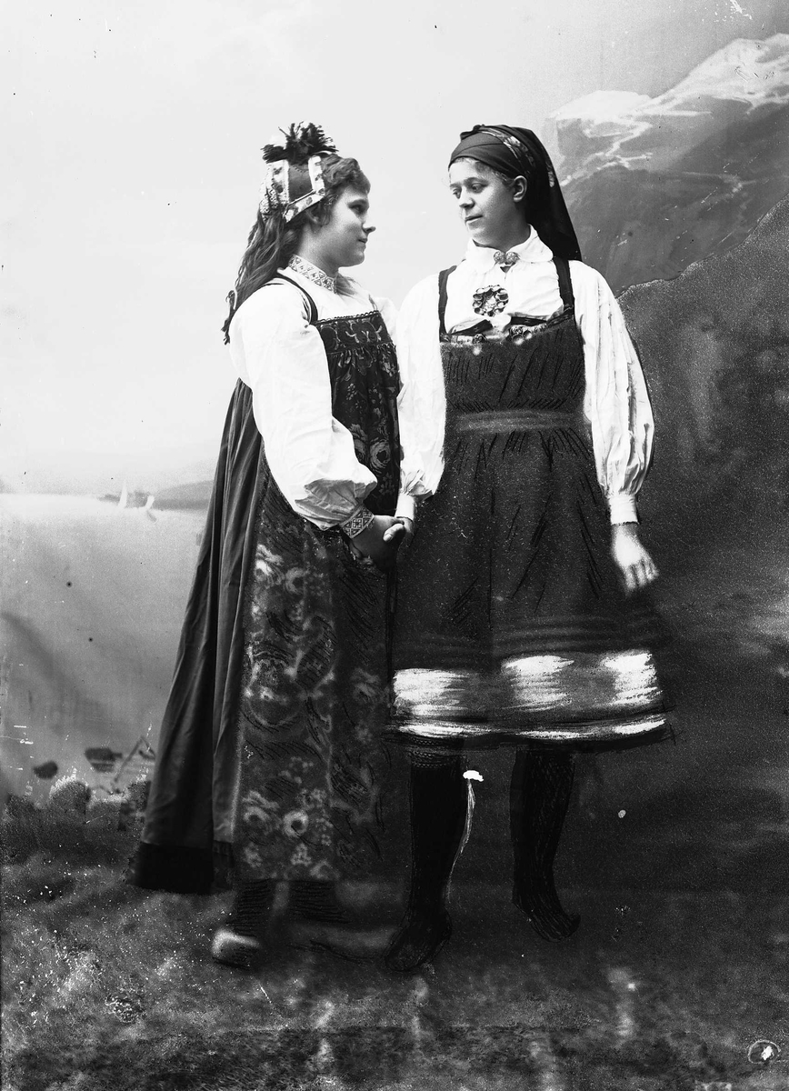 Solveig Lund, Studioportrett av to kvinner i nasjonaldrakter. De ser på hverandre og holder hverandre i hånden. Malt bakgrunn med fjord og fjell.