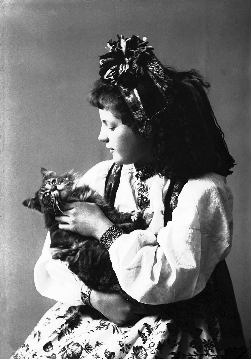 Solveig Lund, 1/2 portrett av kvinne i drakt og skaut, tatt i fotografens studio. Hun sitter med venstre side vendt mot fotografen og koser med en katt hun holder på fanget. Nøytral bakgrunn.