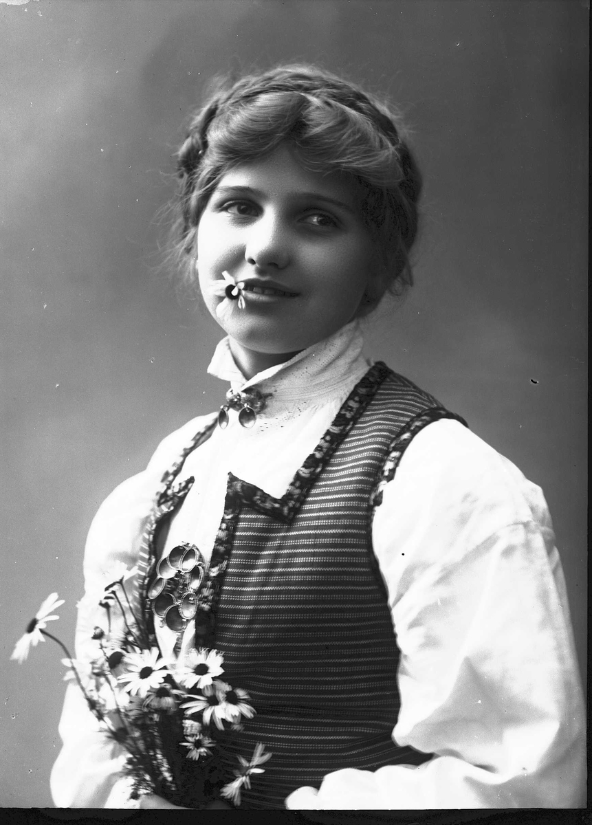 Solveig Lund, Brystportrett av kvinne i drakt med oppsatt hår, fotografert i studio. I munnen holder hun en prestekrage og i hendene en bukett av dem. Kvinnen ser ut av fotografiet. Nøytral bakgrunn.