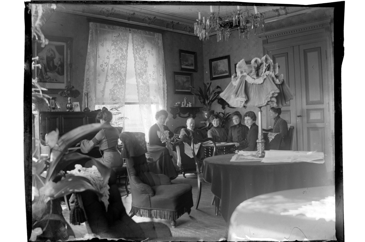 Rolf Sr. Sundt, En mann og seks kvinner, en av dem ved pianoet, i stue på ukjent sted. Fotografert 1903.
