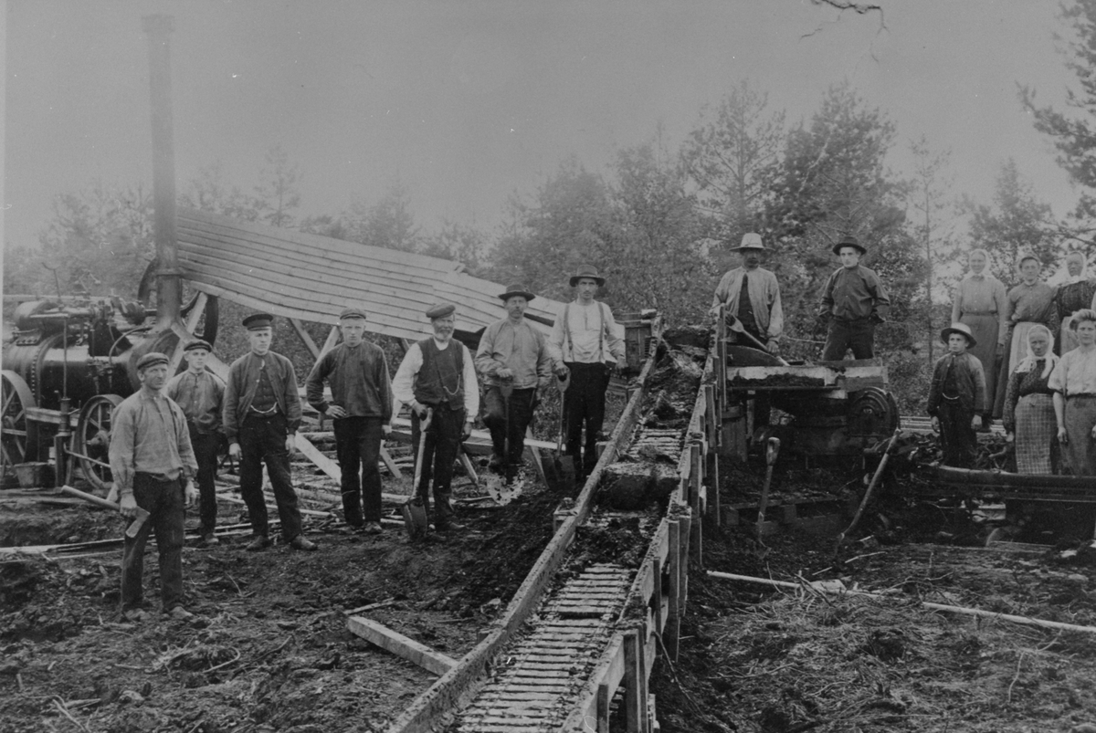 Anno Domkirkeodden, GR: 20, 9 MENN, 1 GUTT, 10 DAMER, ARBEIDSLAG RUNDT TORVMASKIN, DAMPMASKIN, LOKOMOBIL, UKJENT TORVMYR I LØTEN. Kommentar fra Tore Blom, Rubens Maskinhistoriska Samlingar, Götene Sverige. www. rubens. se Denna bild visar en torvtäkt med en Engelsk lokomobil a fabrikatet Ransomes Sims and Jefferies. I…