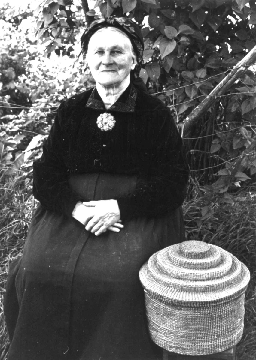 Anna Grostøl, Dølemo Åmli, Aust-Agder 1940. Fru Sigrid Dølemo sitter ved siden av en kurv til å ha silketørklær og sølv i.