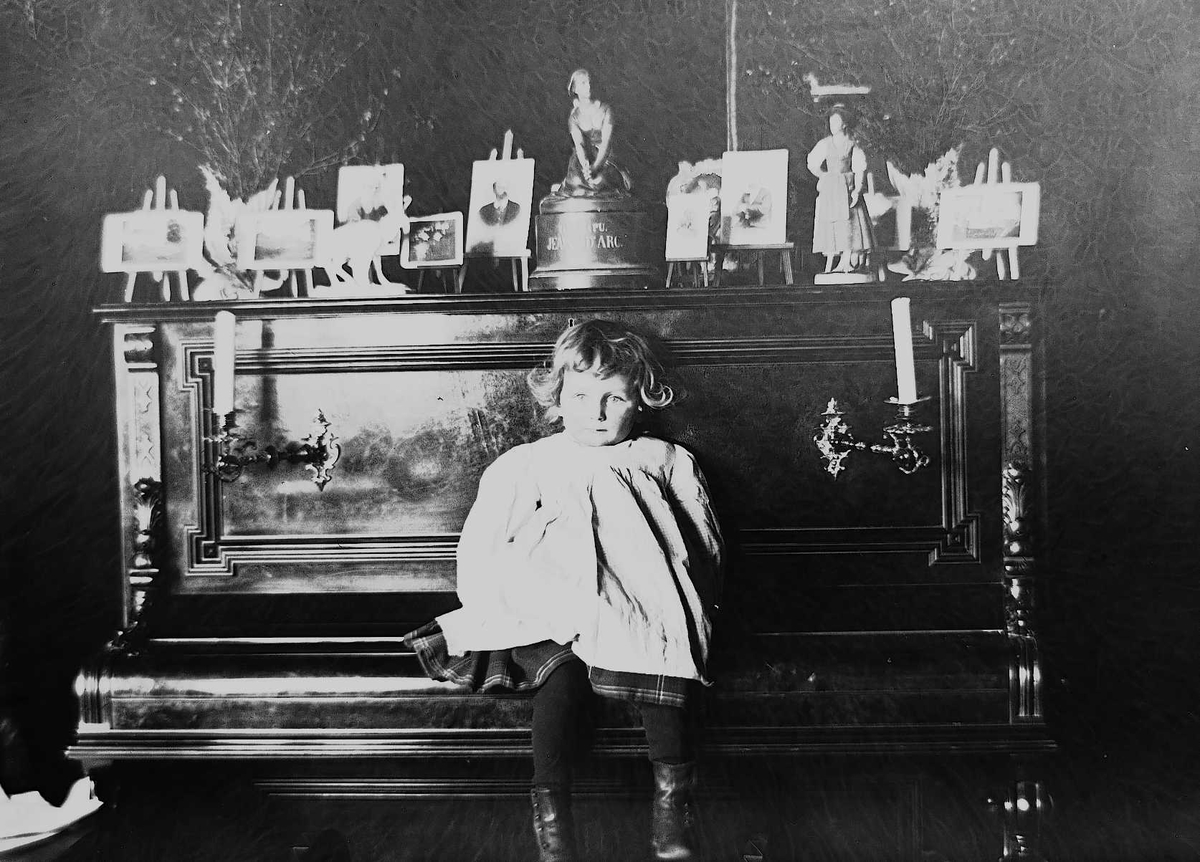 Axel Quinsgaard Wiborg, Gudrun Q. Wiborg sitter på pianolokk i Meltzers gate 9, Oslo, 1899. Fotografier i ramme plassert på pianoet.