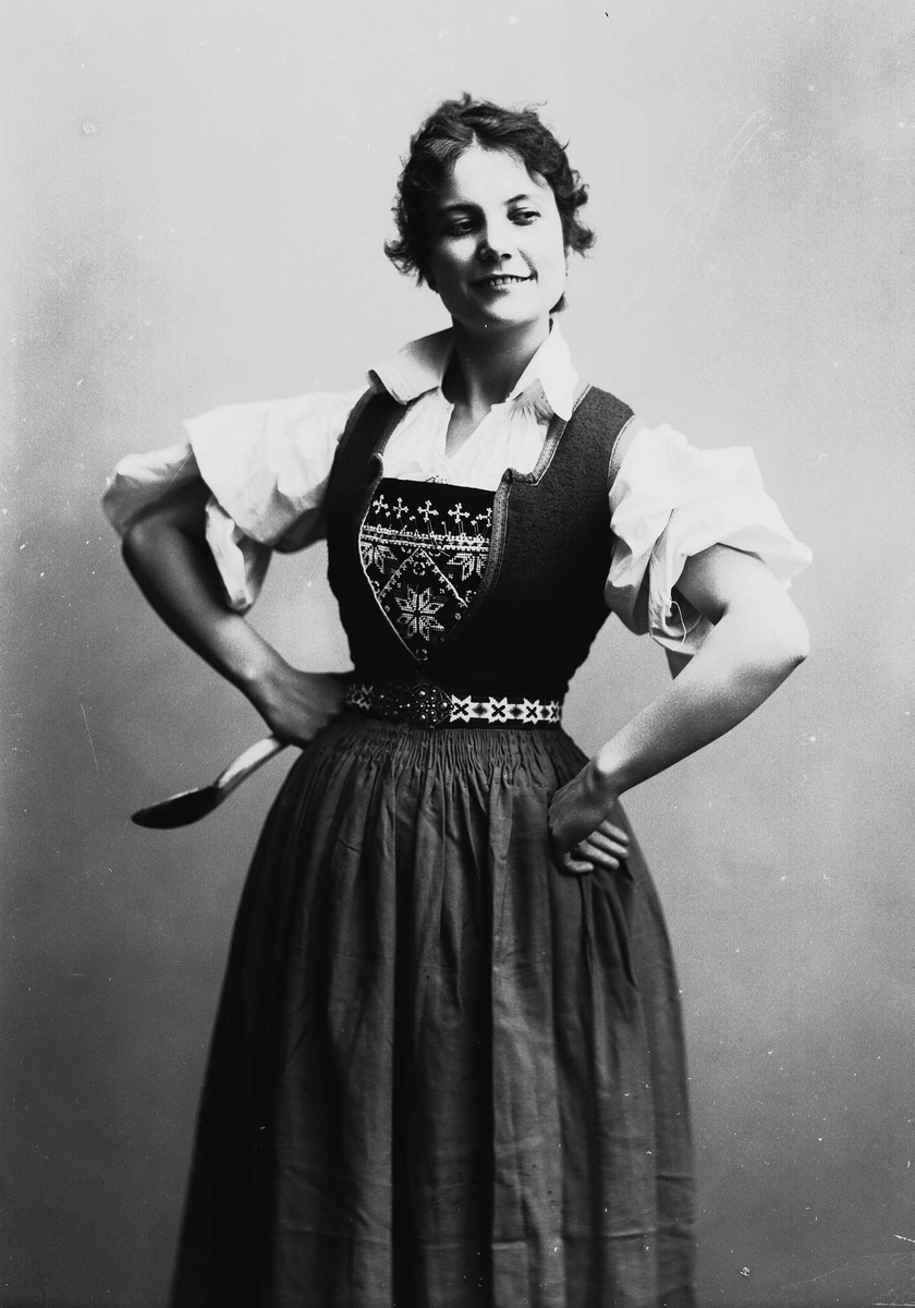 Solveig Lund, 3/4 dels studioportrett av kvinne i drakt. Hun holder hendene på hoften og i den høyre har hun en sleiv.
