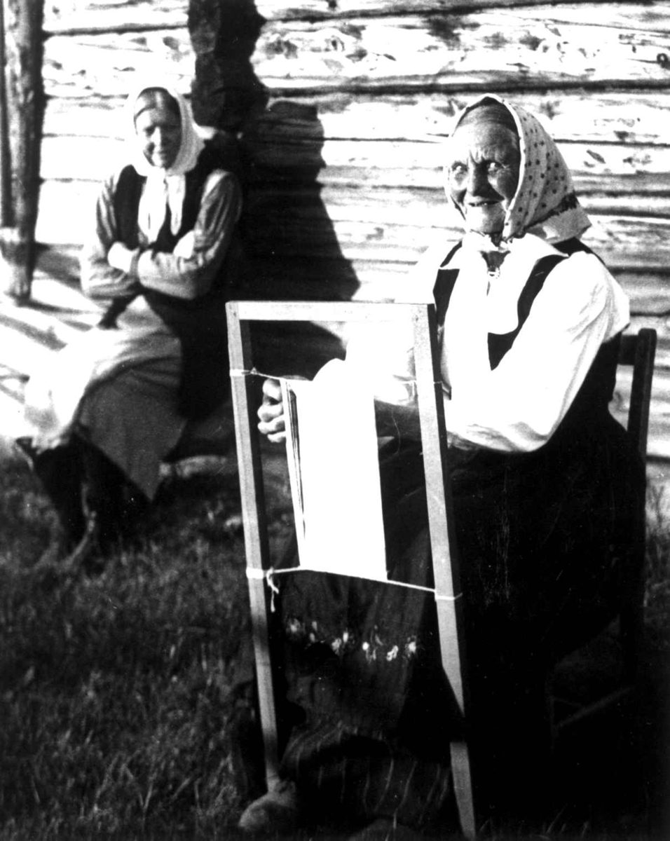 Anna Grostøl, Antagelig Lesja, Oppland 1934. Kvinne i arbeid ved sprrangramme, i bakgrunnen sitter en kvinne ved husveggen og se på.