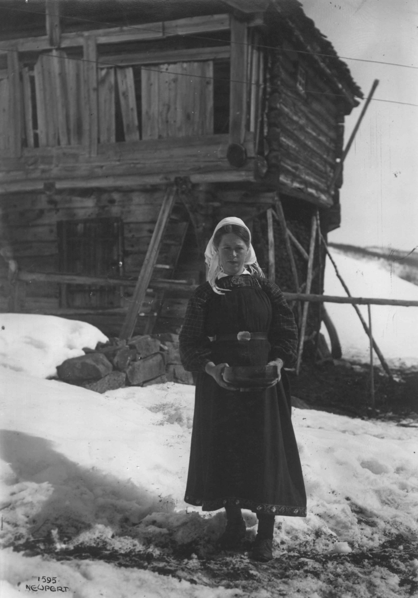 Hermann Christian Neupert, Kvinnedrakt fra Leveld, Ål. Vinterbilde. Kvinne med bolle i hendene. Stående foran et loft (?).