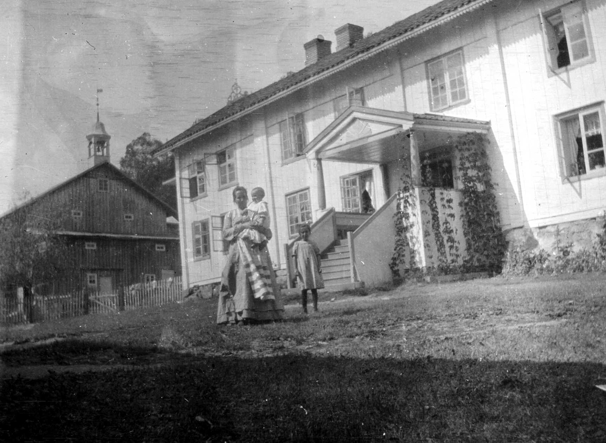Juliane Fredrikke Langberg, Fra gårdstun i Hurum foran våningshus i 2 etasjer med inngangsparti midt på. Bak t.v. stort uthus med klokketårn. Kvinne med to barn i forgrunnen (ukjente). Fra malerinne Juliane Fredrikke Langbergs (1856-1930) samling, tatt ca.1895-1910 med motivkrets slekt og venner samt steder tilknyttet samvær …