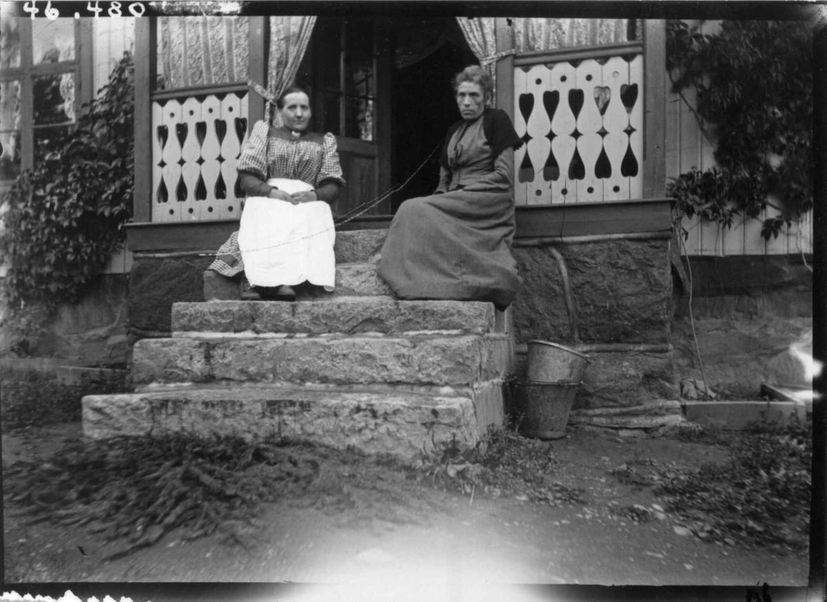 Hermann Christian Neupert, Drakt 1908-10. To kvinner sittende på en trapp. I lange kjoler. I bakgrunnen en veranda.