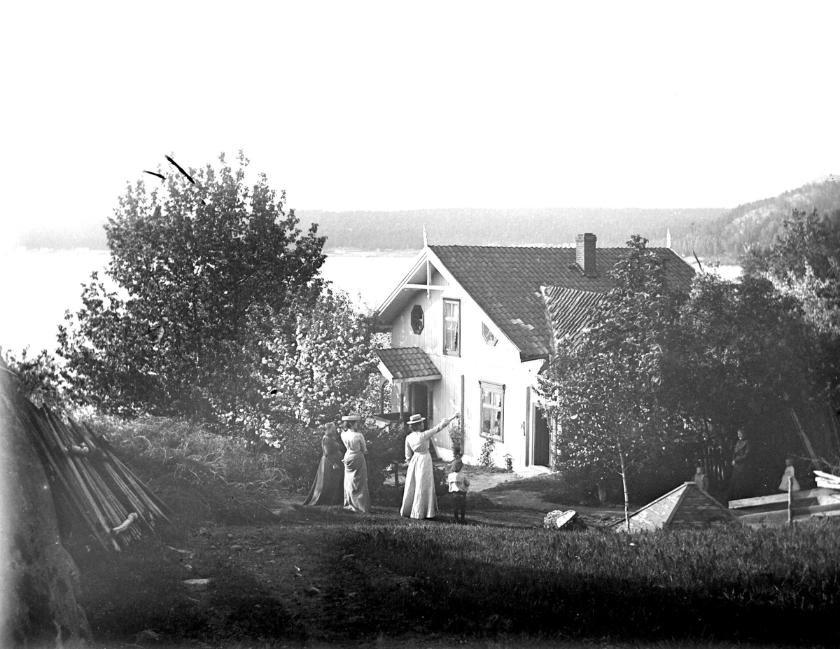 Juliane Fredrikke Langberg, Fra Hurum, persongruppe (kvinner og små barn) utenfor bolighus i sveitserstil (samme hus på NF.15386-029). Brønnhus t.h. Fotografens svoger var prest i Hurum (1897-1904). Fra malerinne Juliane Fredrikke Langbergs (1856-1930) samling, tatt ca.1895-1910 med motivkrets slekt og venner samt steder tilk…