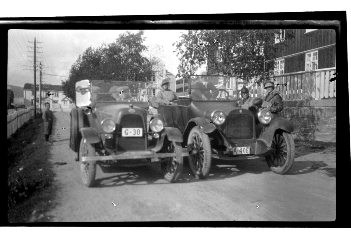 Rolf Sr. Sundt, To biler, eid av henholdsvis Halfdan og Rolf Sundt Sr., med passasjerer under rast på ukjent sted med store trebygninger. Fotografert 1925.
