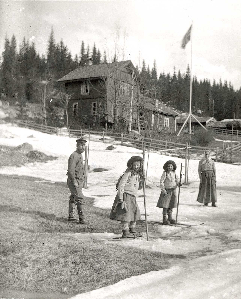 Axel Quinsgaard Wiborg, Persongruppe, Holoa seter, Hadeland, Jevnaker, Oppland, 1906. Pikene på ski er fotografens døtre, Karen og Gudrun Q. Wiborg, sammen med Gulbrand og Lisa (?) foran seterens inngjerdede bolighus og uthus. Del av stereoskopi (se relaterte objekter). Fra serie tatt av iseksportør Axel Quinsgaard Wiborg…