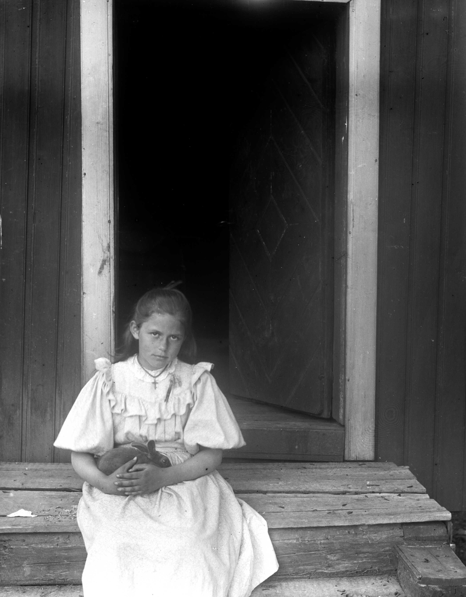 Juliane Fredrikke Langberg, Portrett av jente, Emily Holm (f.1886), fotografens niese, sittende på en trapp foran en halvåpen dør, kledd i lys sommerkjole. Hun har en kanin i fanget. Muligens tatt på kapellangården Bergenhus i Rakkestad (til 1897)eller prestegården i Hurum (fra 1897) , der faren var prest. Fra malerinne Julia…