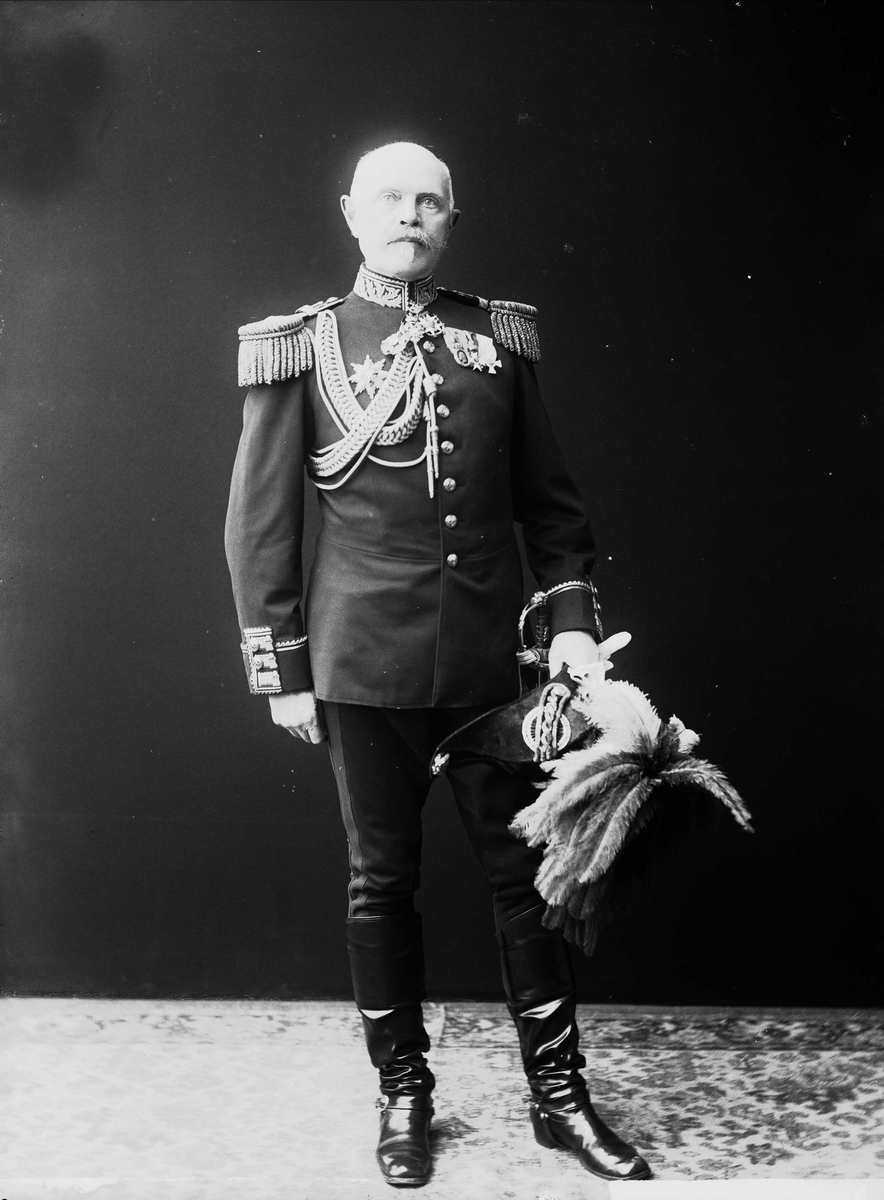 Solveig Lund, Studioportrett av eldre mann i generaluniform stående på teppe foran svart bakgrunn. Holder hatt med fjær i hånden.